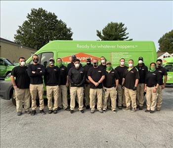 Group of men & women standing in uniforms and masks in front of SERVPRO trucks